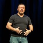 Ricky Gervais standing on stage against black background