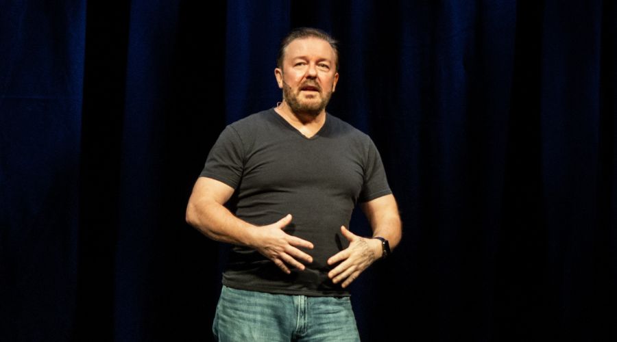 Ricky Gervais standing on stage against black background