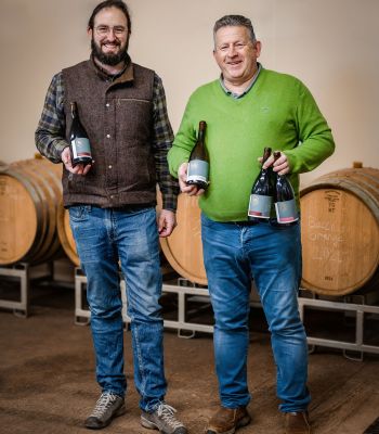 Winemaker Guillaume Lagger and  Beaulieu 58 Wines founder Sandy Booth standing in front of barrels and holding bottles of their wine 